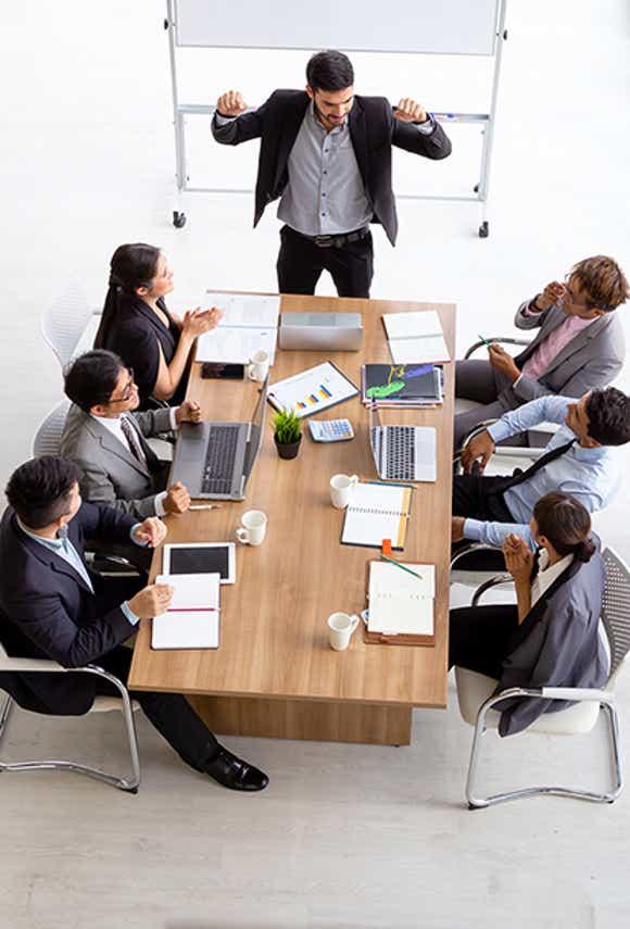 group of sales people gathered around a table looking at a free-standing white board engaged in a meeting lead by a sales leader who uses a structured and simple approach.